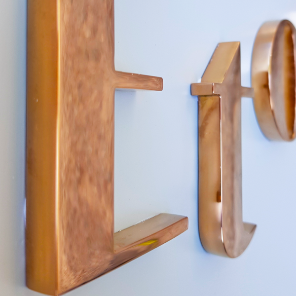Flaggskylt i vit lackerad aluminium med logotyp i individuella profilbokstäver i högblank koppartitanlegerad rostfritt stål – monterad på rund fot och ben i samma material ,
