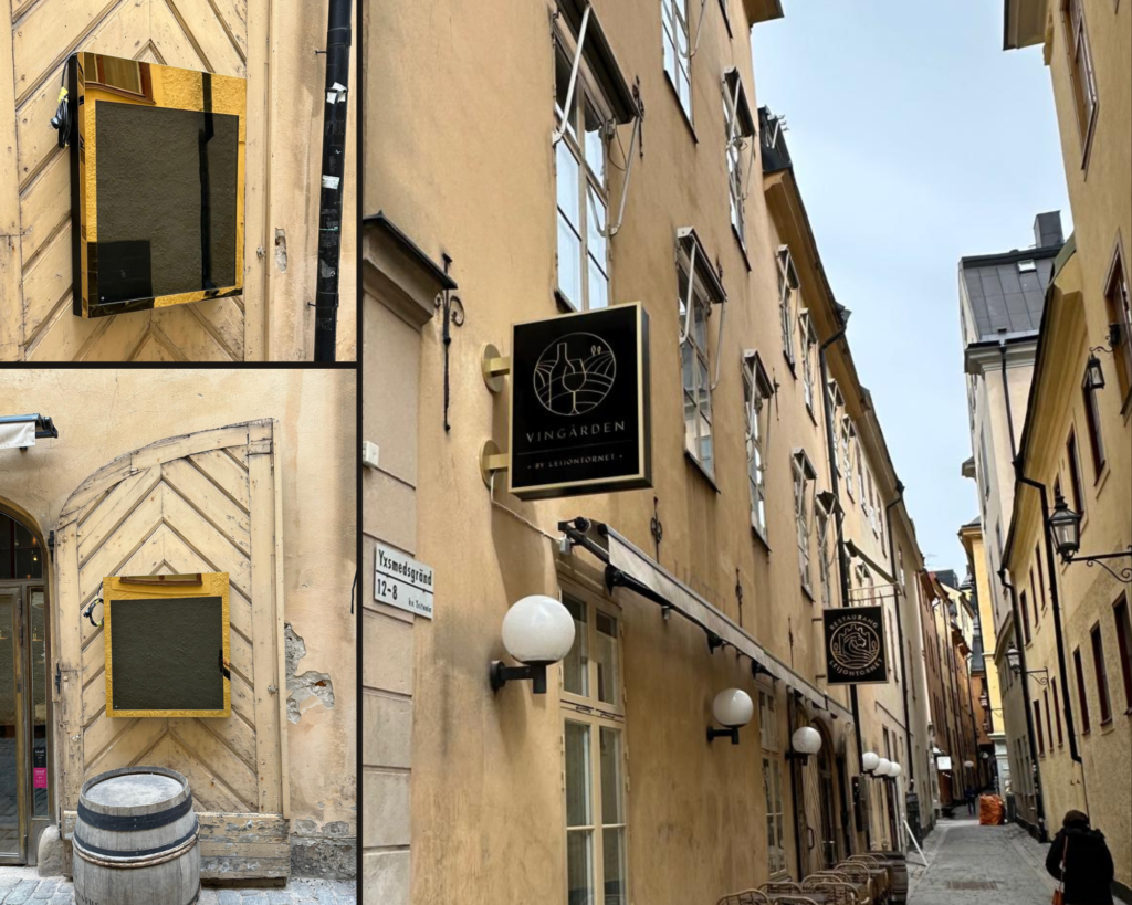 Narrow European street with tan stucco buildings, shop signs, and white globe street lamps along a cobblestone path; outdoor seating visible on the right wall.